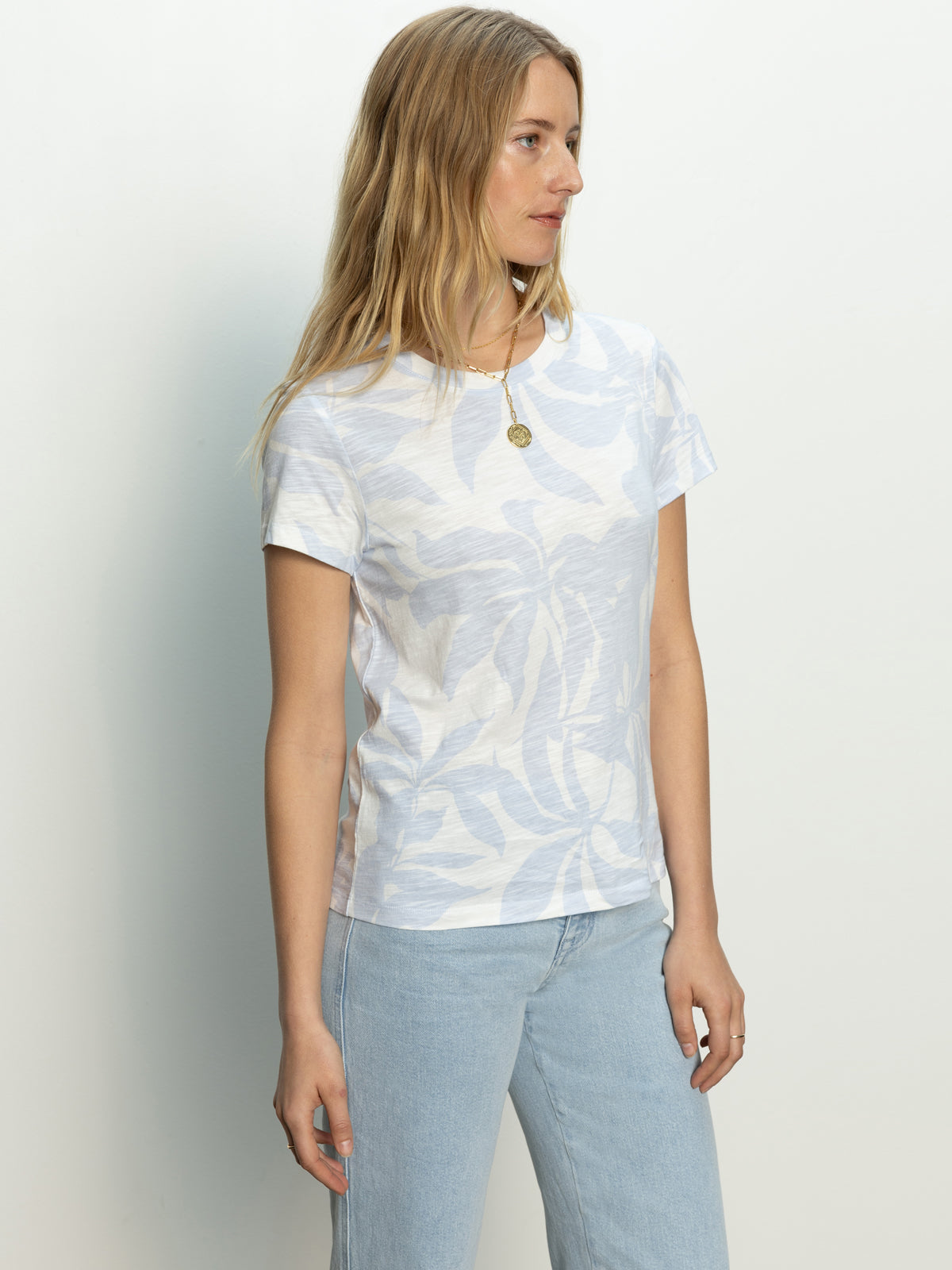 A woman with long blonde hair stands against a plain background, wearing Sanctuary Clothing's "the perfect tee floral bliss" and light blue jeans. She has a calm expression as she looks to the side and wears a round pendant necklace.