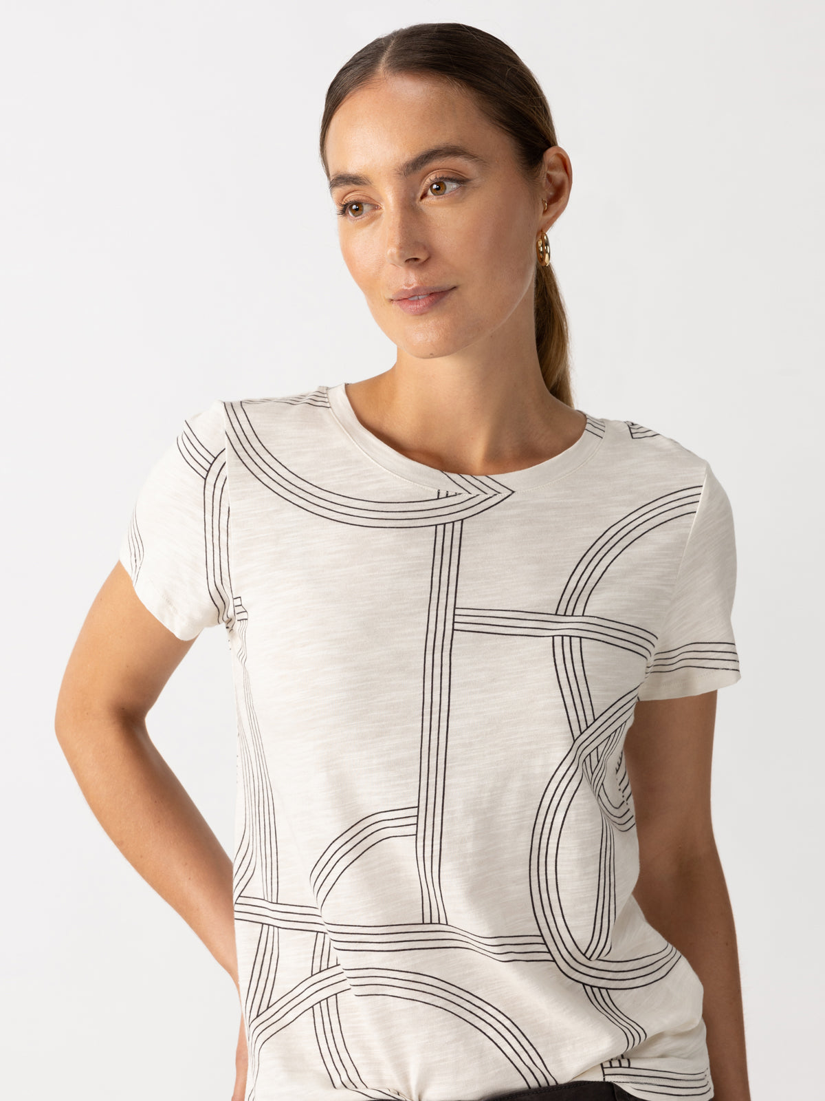 A person with long hair is wearing the Perfect Tee Graphic Lines by Sanctuary Clothing, featuring a black abstract geometric pattern. They have a neutral expression and are looking slightly to the side with one hand on their hip, set against a plain white background.