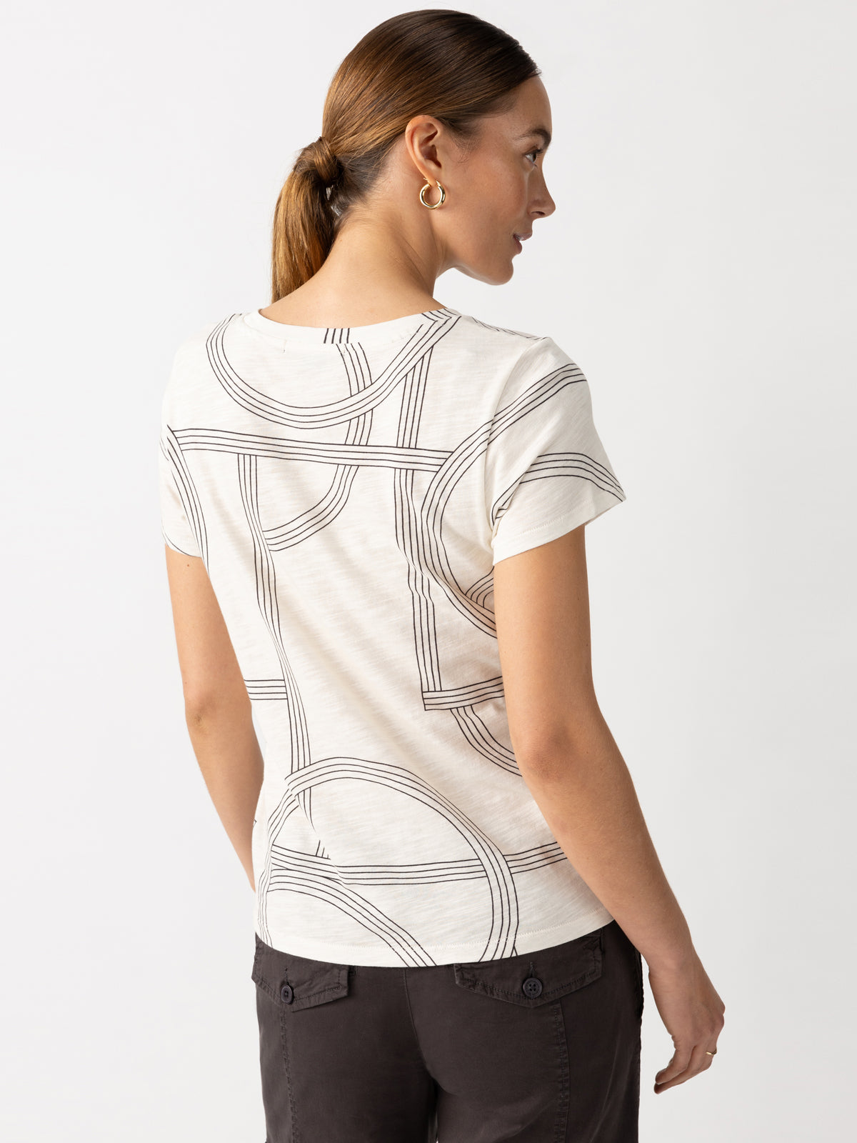 A woman is facing away from the camera, showcasing "The Perfect Tee Graphic Lines" from Sanctuary Clothing. Her medium-length brown hair is tied back, and she complements the look with dark pants and hoop earrings against a plain white background.