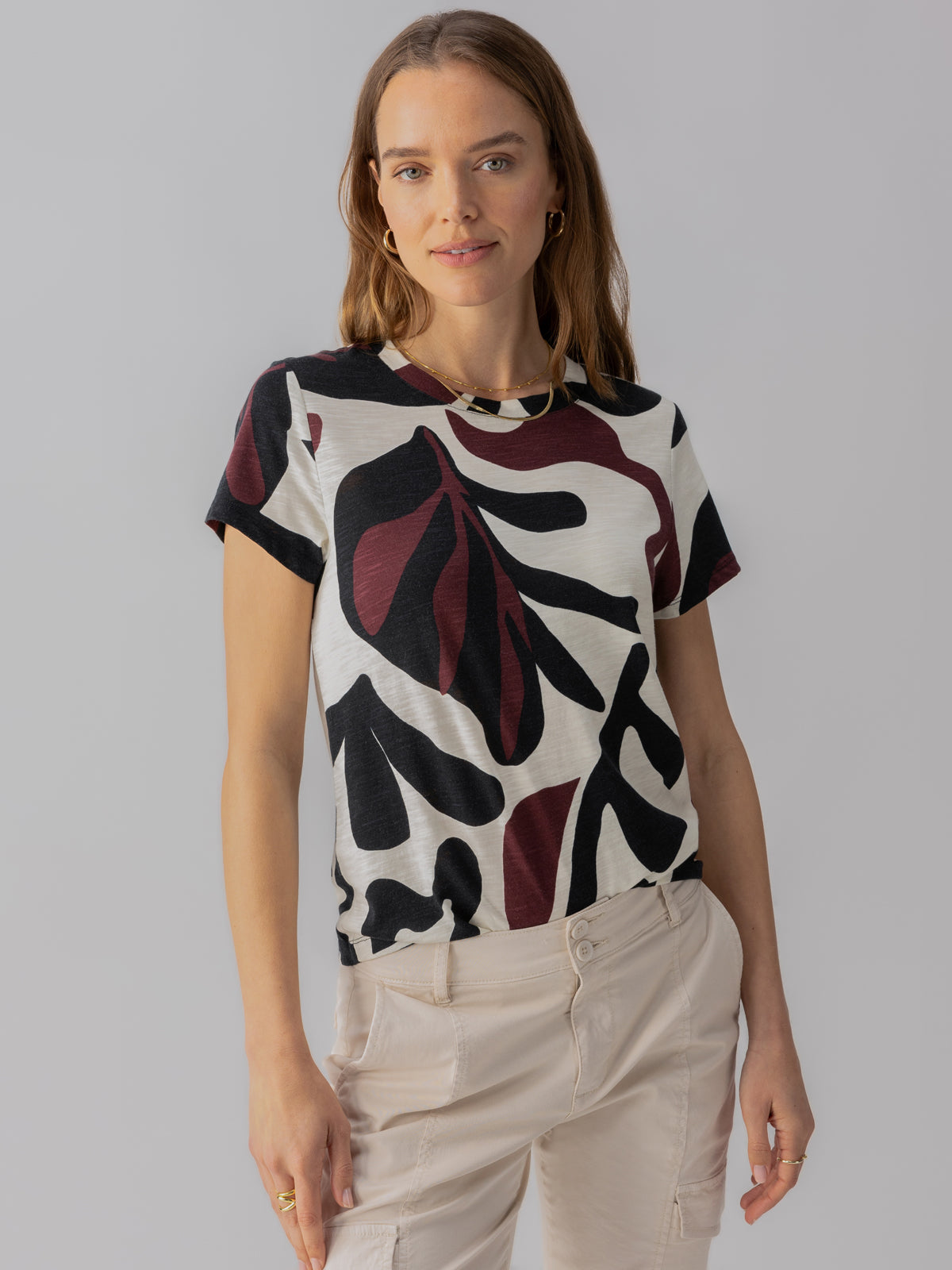 A woman with wavy brown hair is wearing the Perfect Tee Light Mineral by Sanctuary Clothing, featuring an abstract black and dark red leaf pattern. She is standing with a neutral expression against a plain gray background. She also has on light beige pants with visible stitching and pockets.