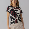 A woman with wavy brown hair is wearing the Perfect Tee Light Mineral by Sanctuary Clothing, featuring an abstract black and dark red leaf pattern. She is standing with a neutral expression against a plain gray background. She also has on light beige pants with visible stitching and pockets.