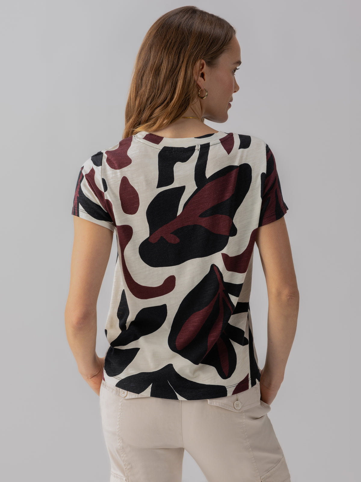 A person with long hair is seen from behind, wearing Sanctuary Clothing's short-sleeved, white shirt called The Perfect Tee Light Mineral, featuring an abstract maroon and black leaf pattern, paired with light-colored pants. The background is plain and grey.