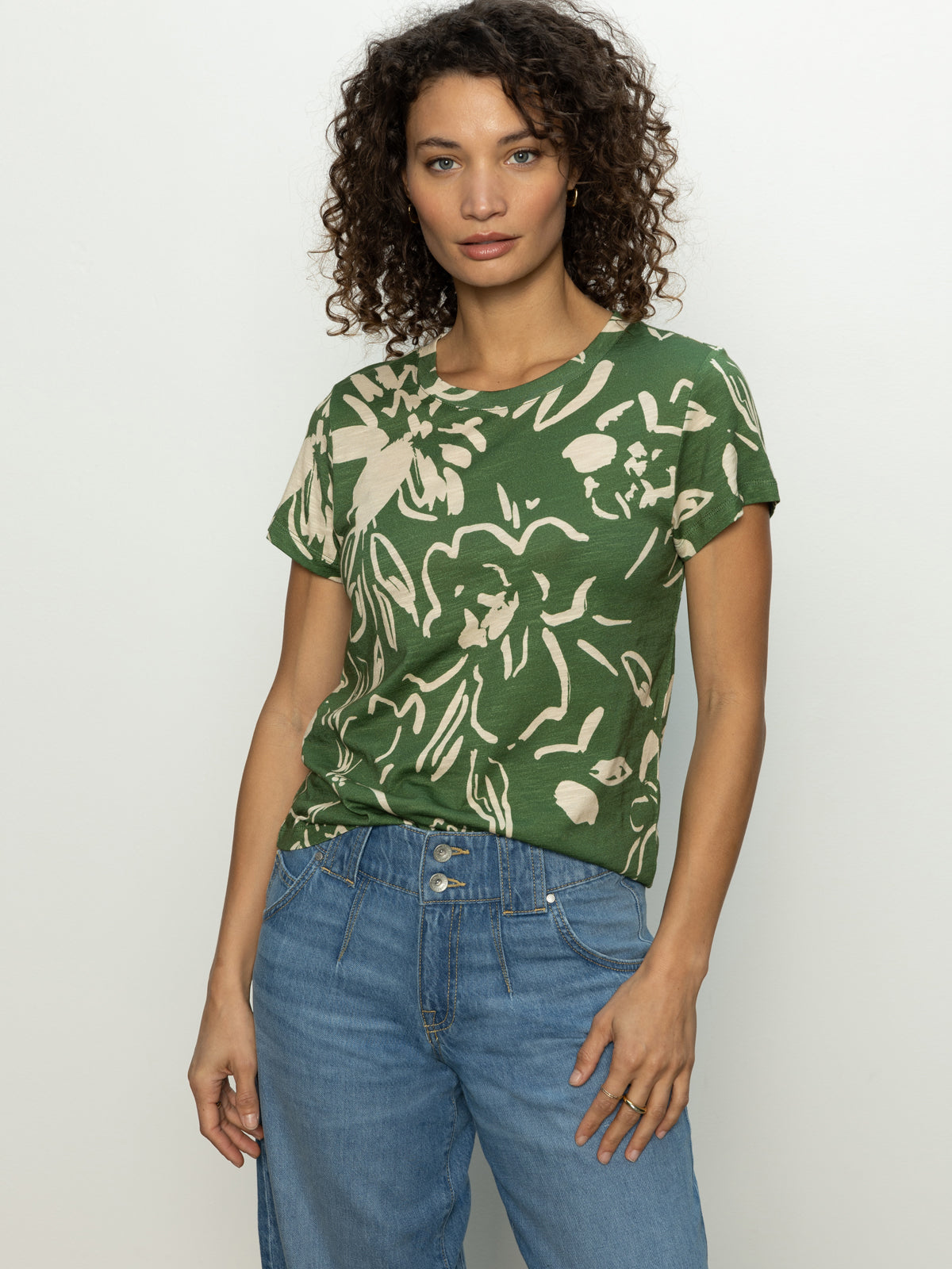 A person with curly hair, wearing Sanctuary Clothing's "the perfect tee lush harmony" floral-patterned green shirt and blue jeans, stands against a plain white background.