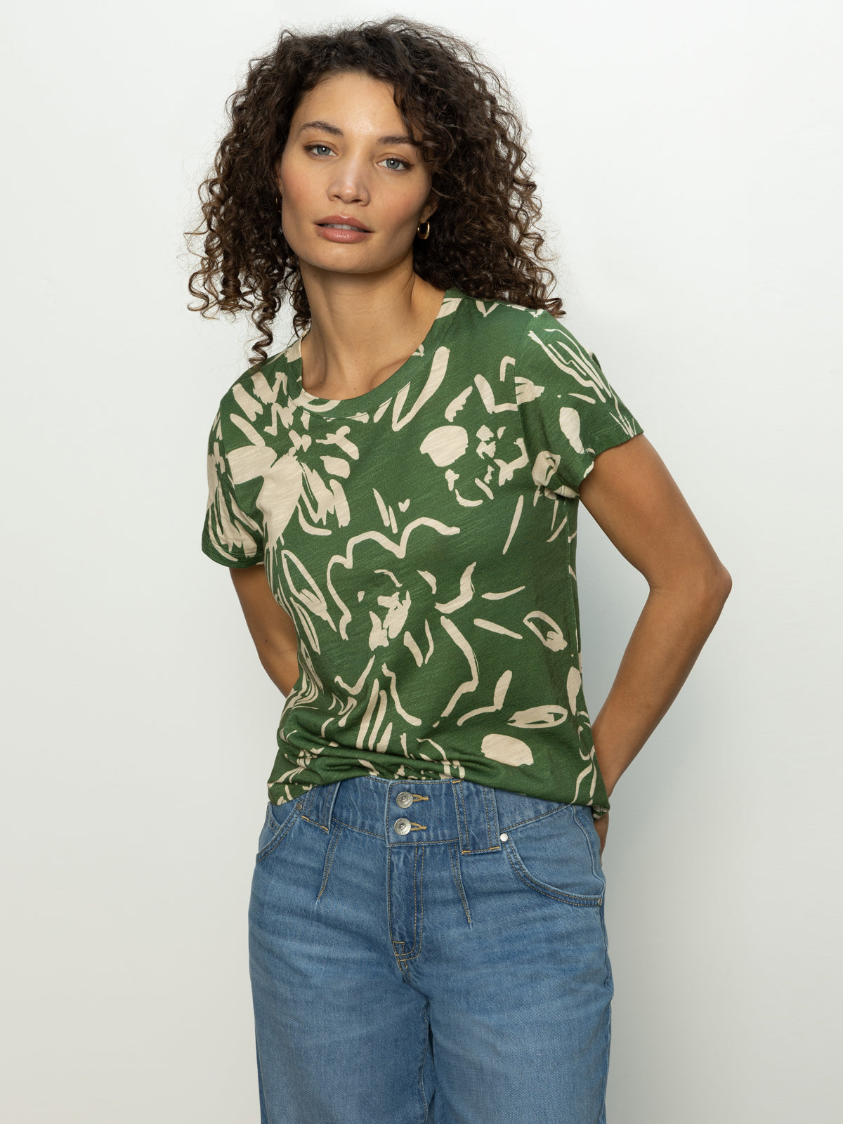 A person with curly hair is wearing the Perfect Tee Lush Harmony by Sanctuary Clothing, featuring a green floral pattern. They're styled in blue jeans and posing with hands in pockets against a plain background.