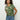 A person with curly hair is wearing the Perfect Tee Lush Harmony by Sanctuary Clothing, featuring a green floral pattern. They're styled in blue jeans and posing with hands in pockets against a plain background.