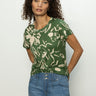 A person with curly hair is wearing the Perfect Tee Lush Harmony by Sanctuary Clothing, featuring a green floral pattern. They're styled in blue jeans and posing with hands in pockets against a plain background.