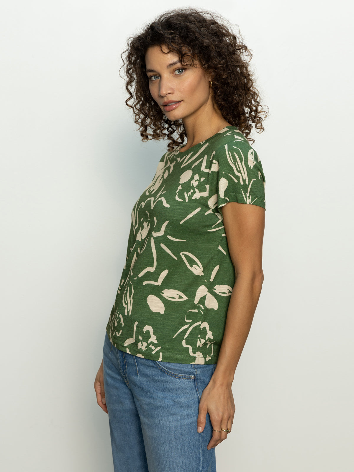 A person with curly hair is wearing Sanctuary Clothing's "the perfect tee lush harmony," featuring a beige floral pattern on green, and is standing in profile against a plain background. They're paired with blue jeans.