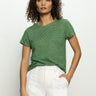 A woman with curly hair wears Sanctuary Clothing's "the perfect tee palm chalk stripe," a green and black striped t-shirt, paired with white pants, set against a plain background.