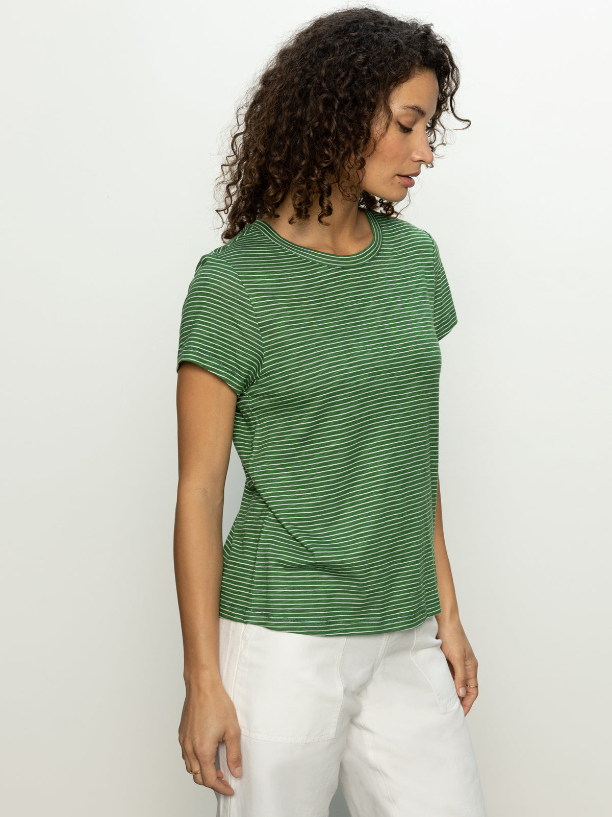 A woman with curly hair is wearing Sanctuary Clothing's "the perfect tee palm chalk stripe" and white pants, standing against a plain background.