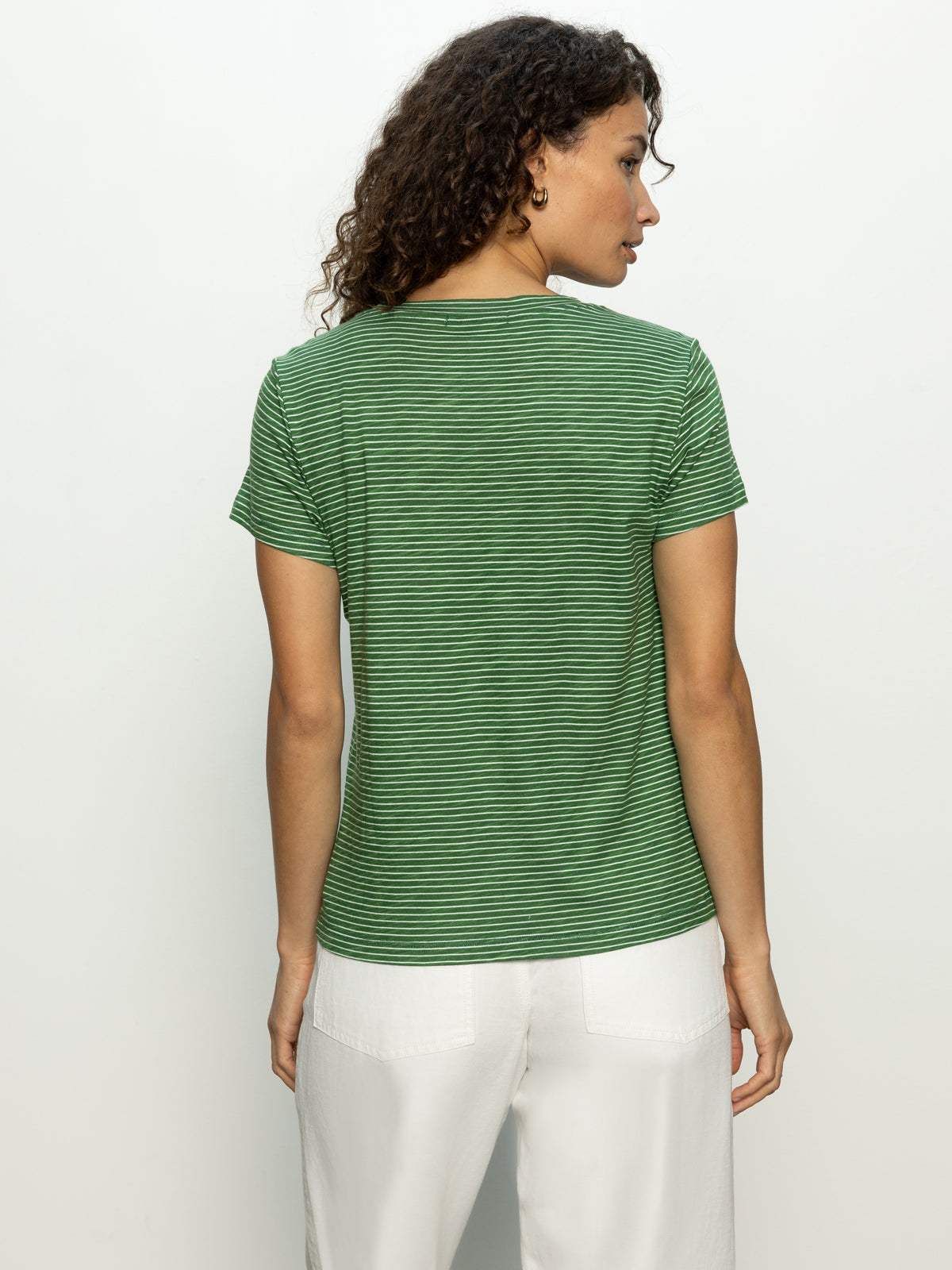 A person with curly hair is facing away, wearing Sanctuary Clothing's "The Perfect Tee" in palm chalk stripe with white pants, set against a plain, light background.