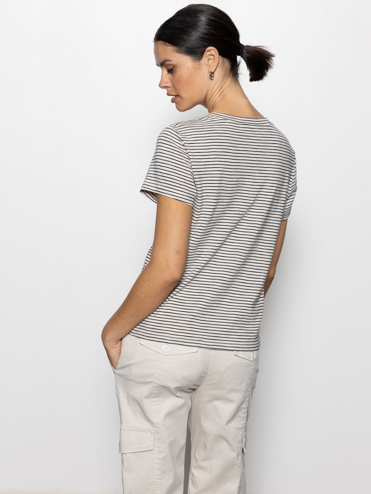 A person wearing the Sanctuary Clothing "the perfect tee pumice stone black stripe" stands with their back to the camera, head turned to the side. Their hands are in their pockets against a plain white background, paired with light-colored pants.