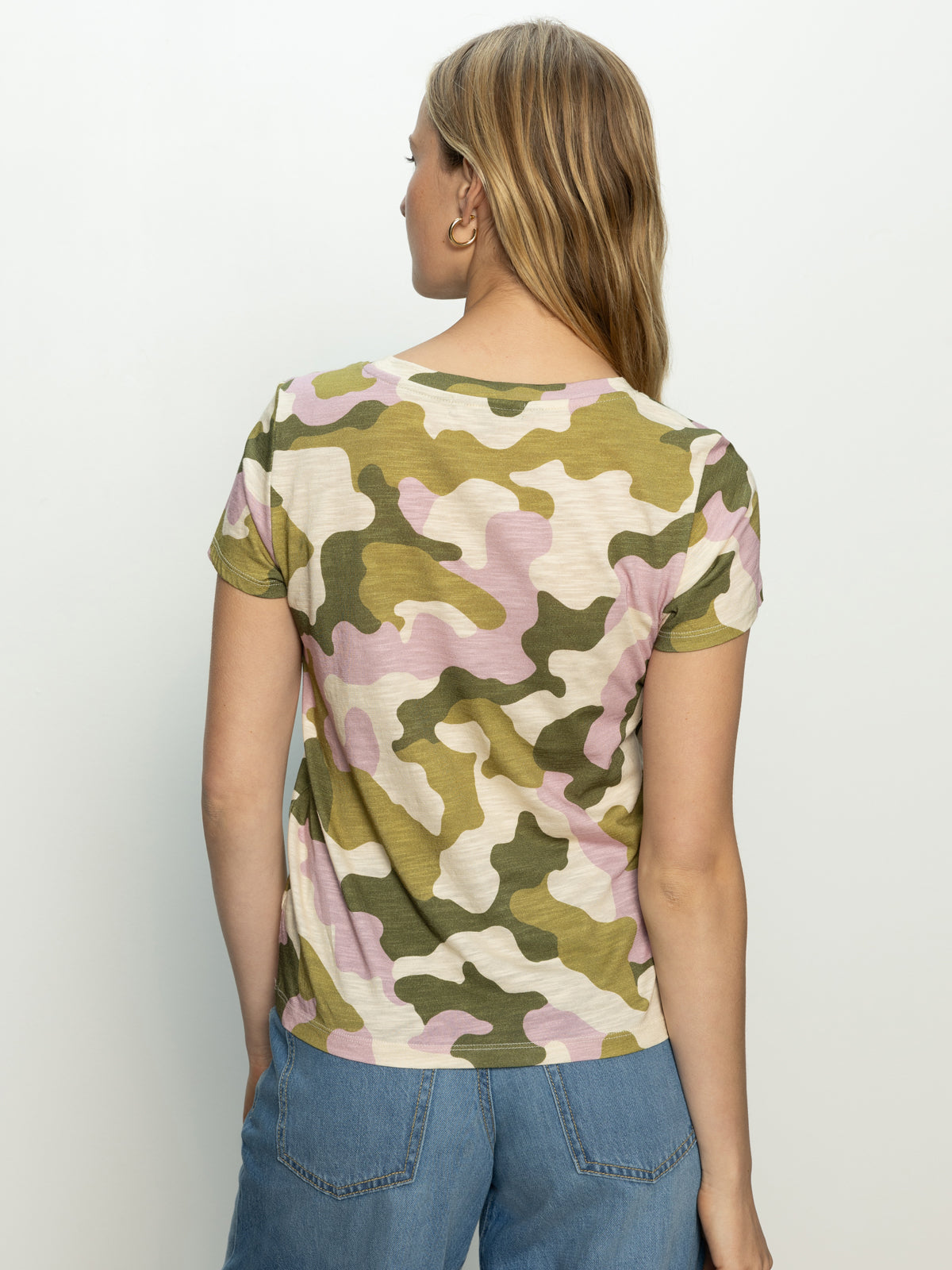 A person with long hair, seen from behind, is wearing Sanctuary Clothing's "the perfect tee precious camo" T-shirt in shades of green, pink, and beige, paired with blue jeans against a plain light gray background.