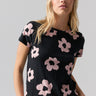 A person with long blonde hair is standing against a plain background. They are wearing The Perfect Tee Rose Smoke Flower Pop from Sanctuary Clothing, featuring large, pale pink flower prints. Their right hand is resting in their pocket, and they have a neutral expression on their face.