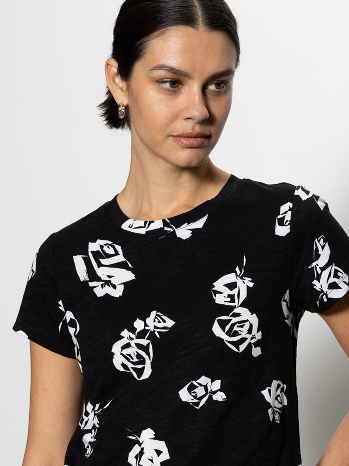 A woman wearing "The Perfect Tee Rose Pop" by Sanctuary Clothing, with white abstract floral patterns over a black background, is looking slightly to the side. Her dark hair is pulled back, and she stands against a plain white background.