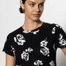 A woman wearing "The Perfect Tee Rose Pop" by Sanctuary Clothing, with white abstract floral patterns over a black background, is looking slightly to the side. Her dark hair is pulled back, and she stands against a plain white background.