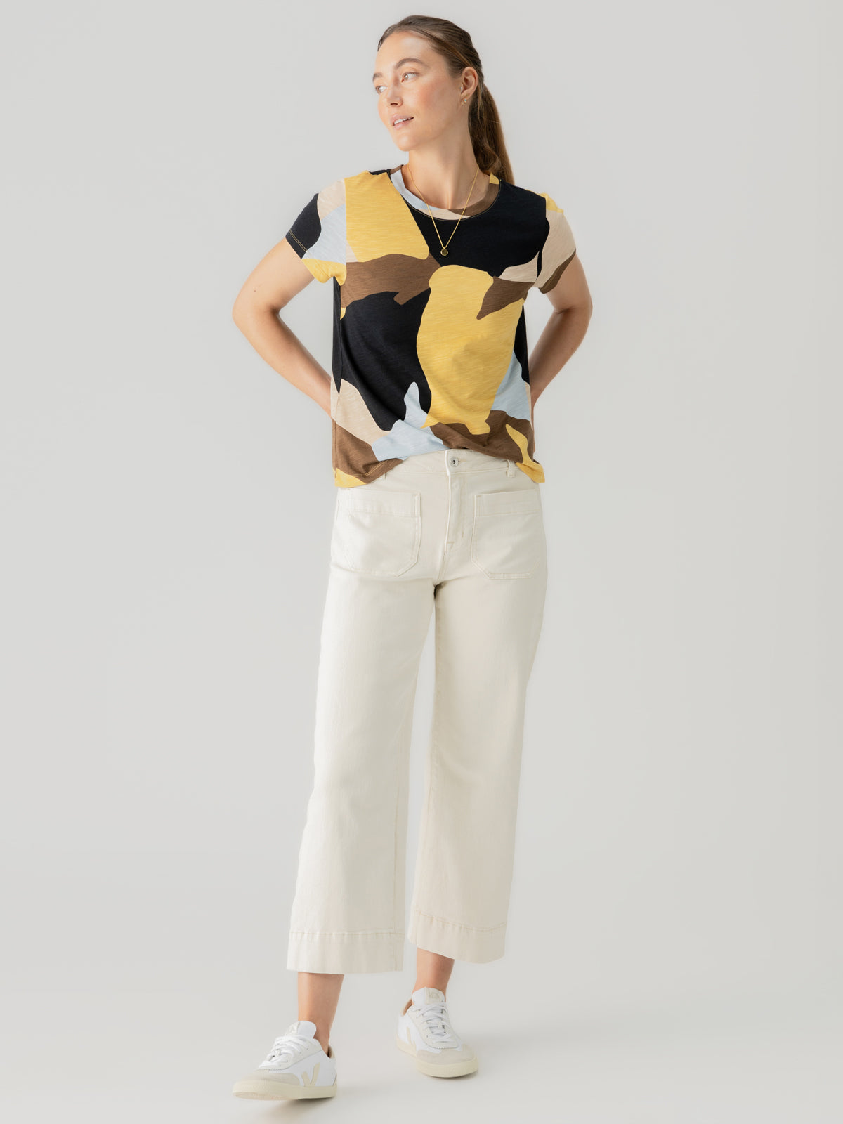 A woman stands in a relaxed pose, wearing Sanctuary Clothing's "The Perfect Tee Sky Escape," with its colorful abstract pattern. She pairs it with white wide-leg pants and white sneakers. Her hands are on her hips as she looks off to the side, her hair pulled back in a ponytail. The background is plain and light-colored.