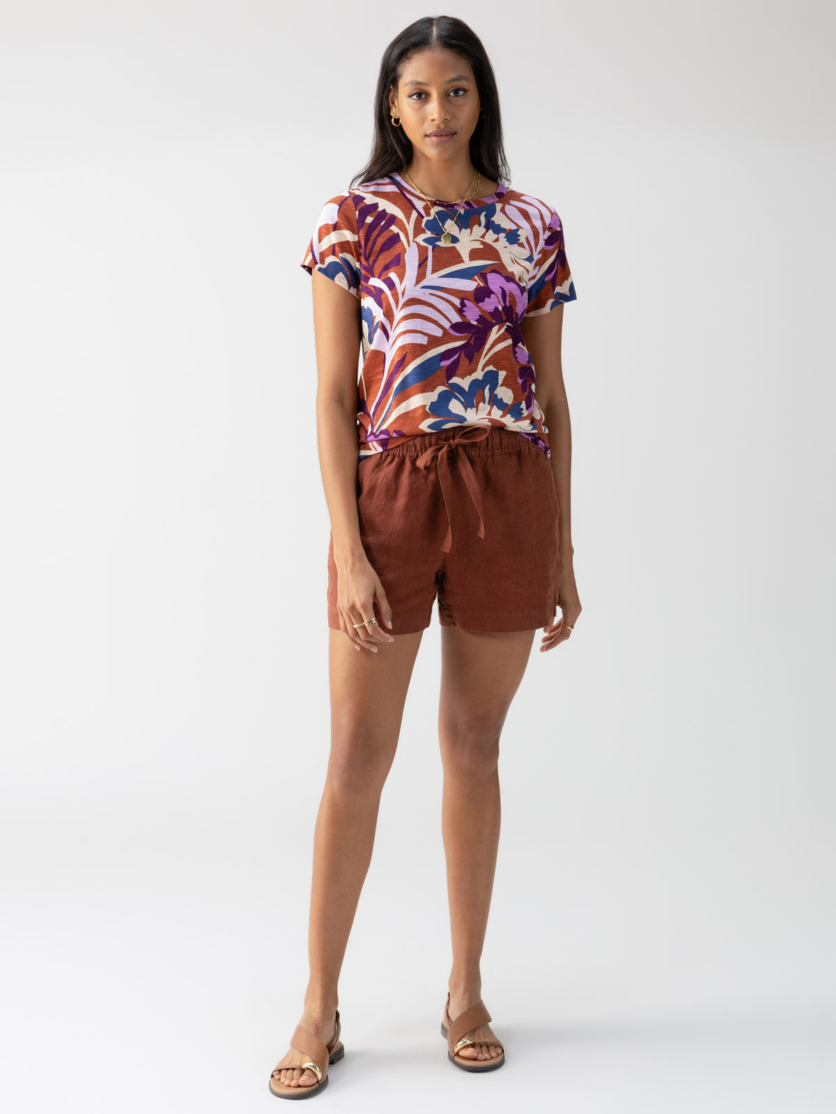 A woman stands against a light grey background. She is wearing Sanctuary Clothing's "The Perfect Tee South Palm," a colorful floral-print T-shirt with shades of purple, blue, and orange, paired with rust-colored shorts tied at the waist. She completes the outfit with brown sandals.