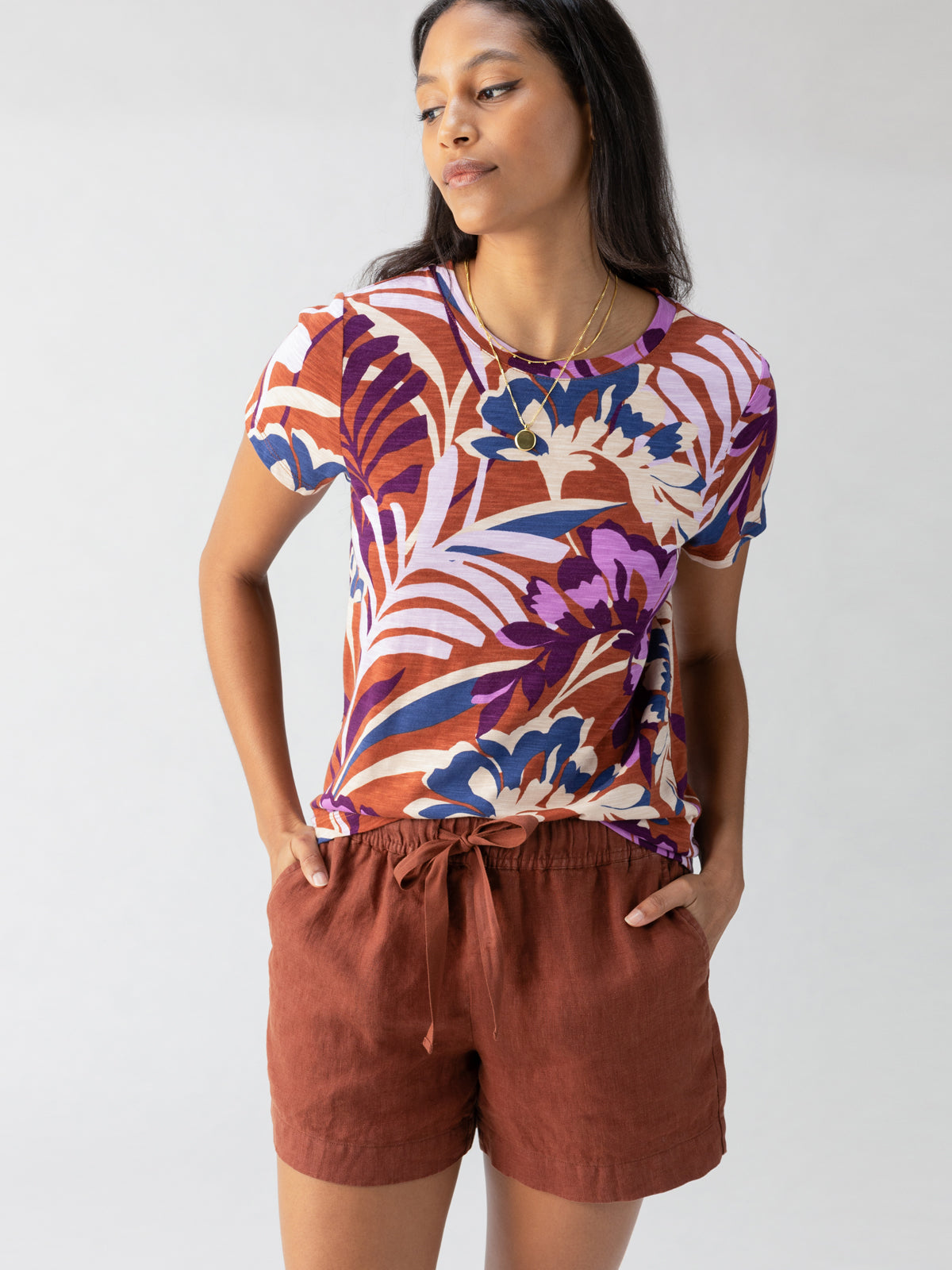 A person with long hair stands confidently, wearing Sanctuary Clothing’s The Perfect Tee South Palm short-sleeved shirt and rust-colored shorts. They have their hands on their hips and are looking slightly to the side. The background is plain and light-colored.