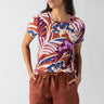 A person with long hair stands confidently, wearing Sanctuary Clothing’s The Perfect Tee South Palm short-sleeved shirt and rust-colored shorts. They have their hands on their hips and are looking slightly to the side. The background is plain and light-colored.