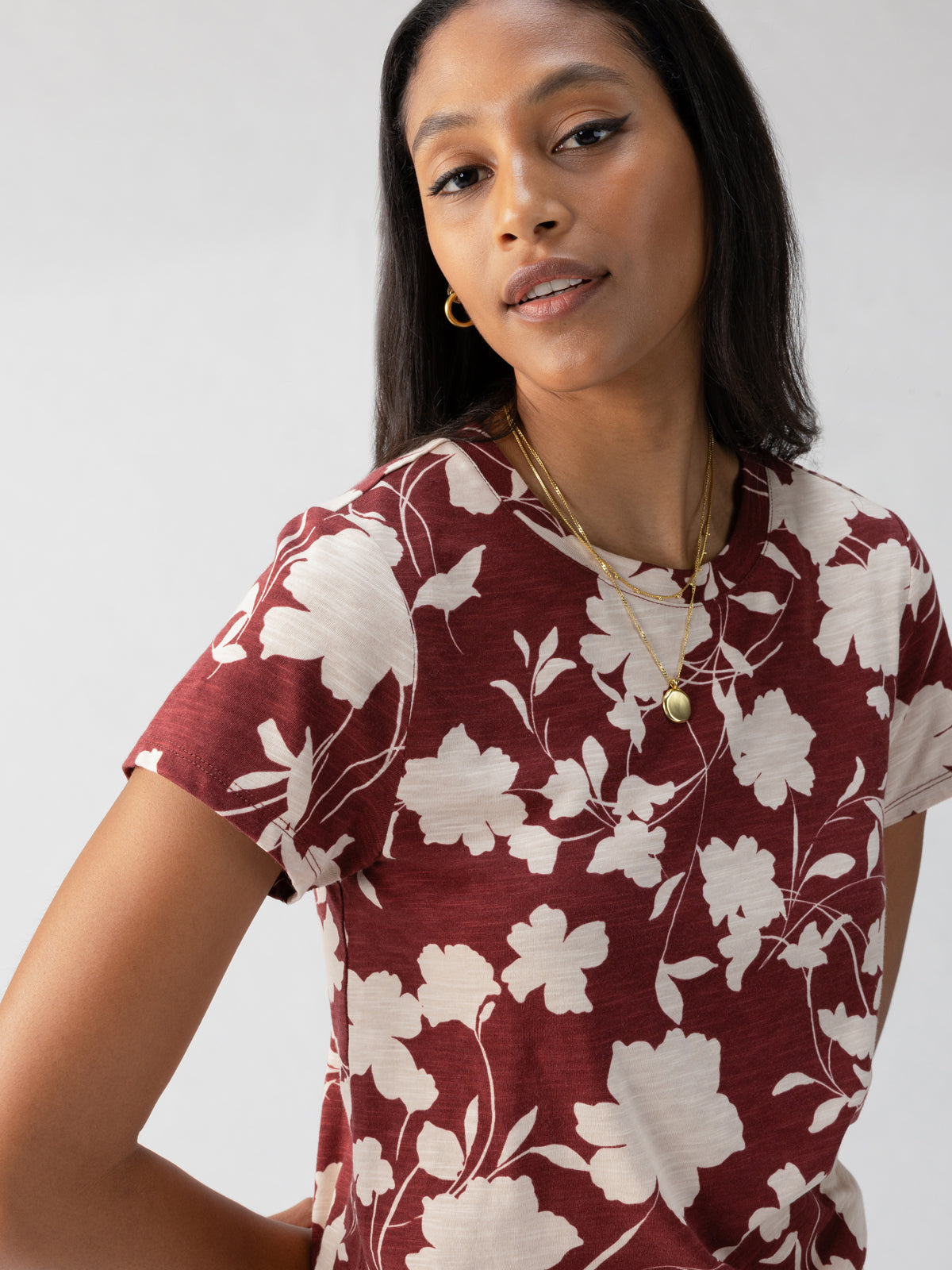 A woman with long dark hair is seen wearing Sanctuary Clothing's "The Perfect Tee Warm Vista," a maroon shirt adorned with cream-colored floral patterns. She accessorizes with gold hoop earrings and layered gold necklaces, gazing slightly off-camera with a neutral expression. The background is plain and light-colored.