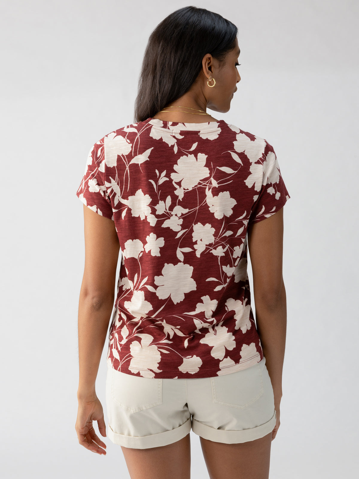 A woman is facing away from the camera, wearing "The Perfect Tee Warm Vista" by Sanctuary Clothing. This maroon short-sleeved shirt features a white floral pattern and is paired with light-colored shorts. She has long, dark hair and is standing against a light gray background.