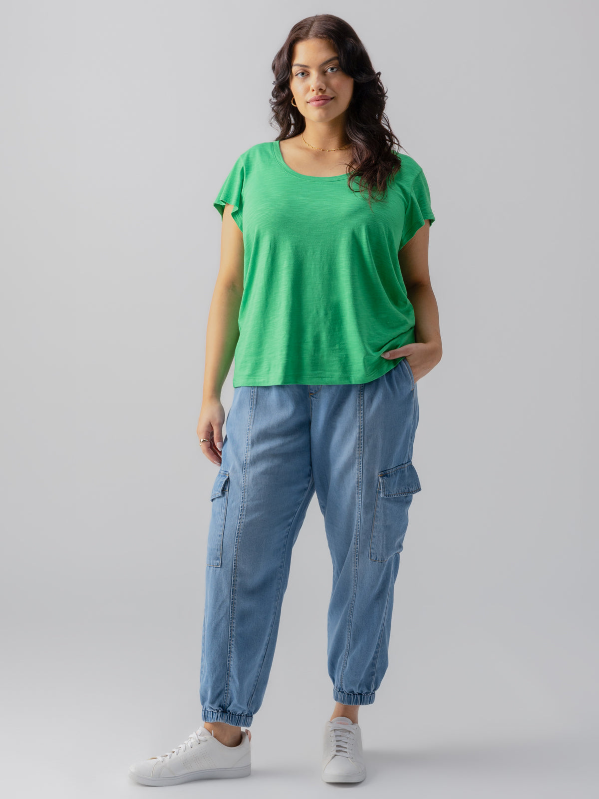 A person stands against a plain grey background, sporting the Sanctuary Clothing's West Side Tee in Green Goddess from their Inclusive Collection and casual light blue cargo pants. They have wavy dark hair, white sneakers, one hand in their pocket, and a slight, confident smile on their face.