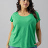 A person with long, dark, wavy hair is wearing the bright green short-sleeve West Side Tee from the Green Goddess Inclusive Collection by Sanctuary Clothing and light blue jeans. They are posing with one hand in the pocket of their jeans against a plain gray background, looking slightly to the side.