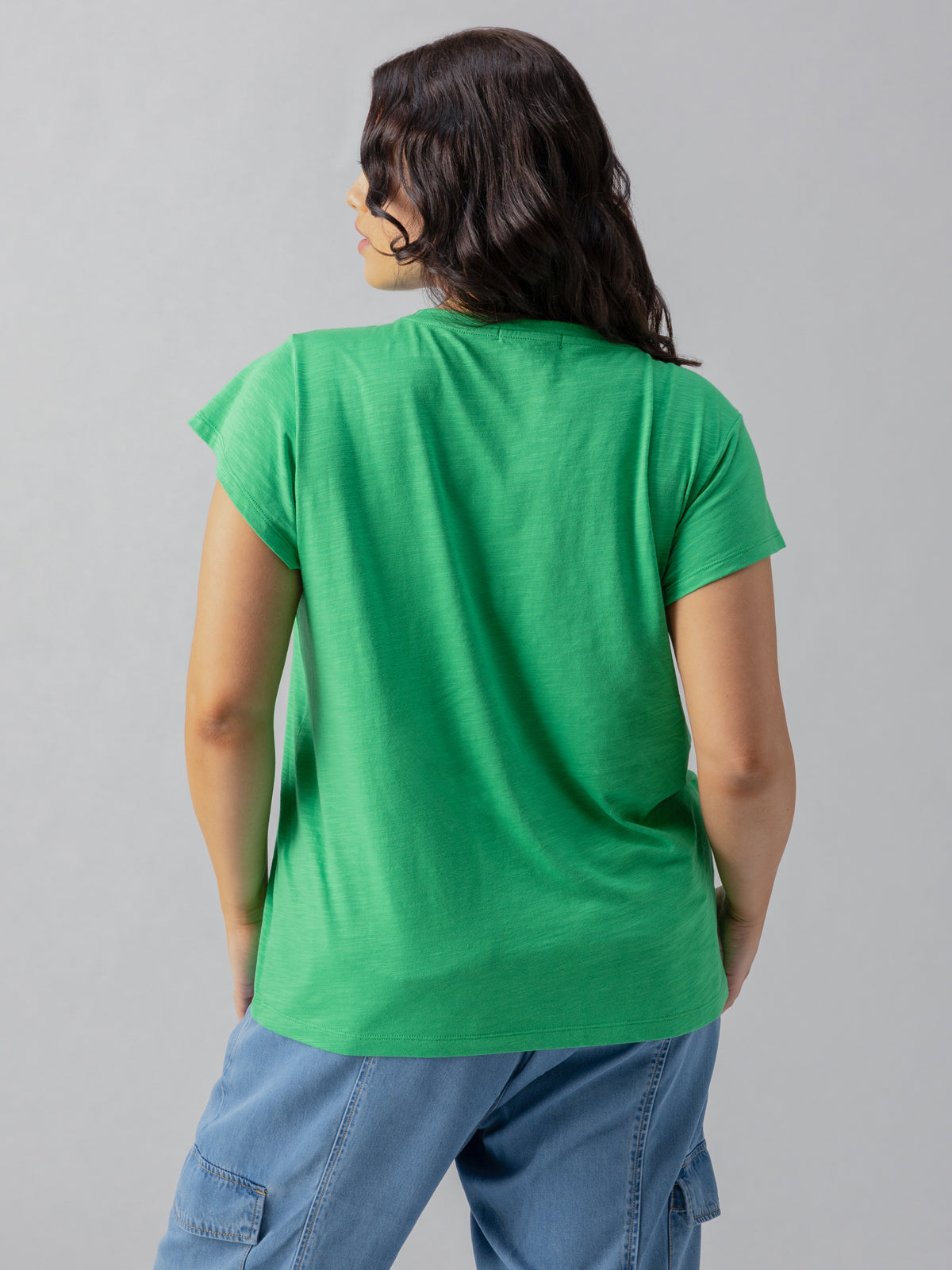 A person with dark hair stands with their back to the camera, wearing the Sanctuary Clothing West Side Tee Green Goddess from the Inclusive Collection and light blue pants with cargo pockets. The background is plain and light-colored.