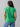 A person with dark hair stands with their back to the camera, wearing the Sanctuary Clothing West Side Tee Green Goddess from the Inclusive Collection and light blue pants with cargo pockets. The background is plain and light-colored.