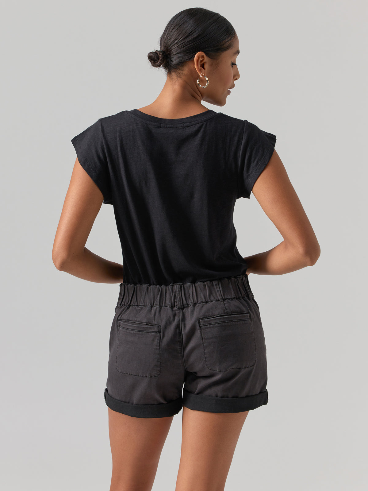 A person with dark hair is shown from behind, wearing the Sanctuary Clothing West Side Tee in black paired with dark gray shorts featuring pockets. The hair is tied back in a low, neat bun, and the person is adorned with gold hoop earrings against a plain light gray background.