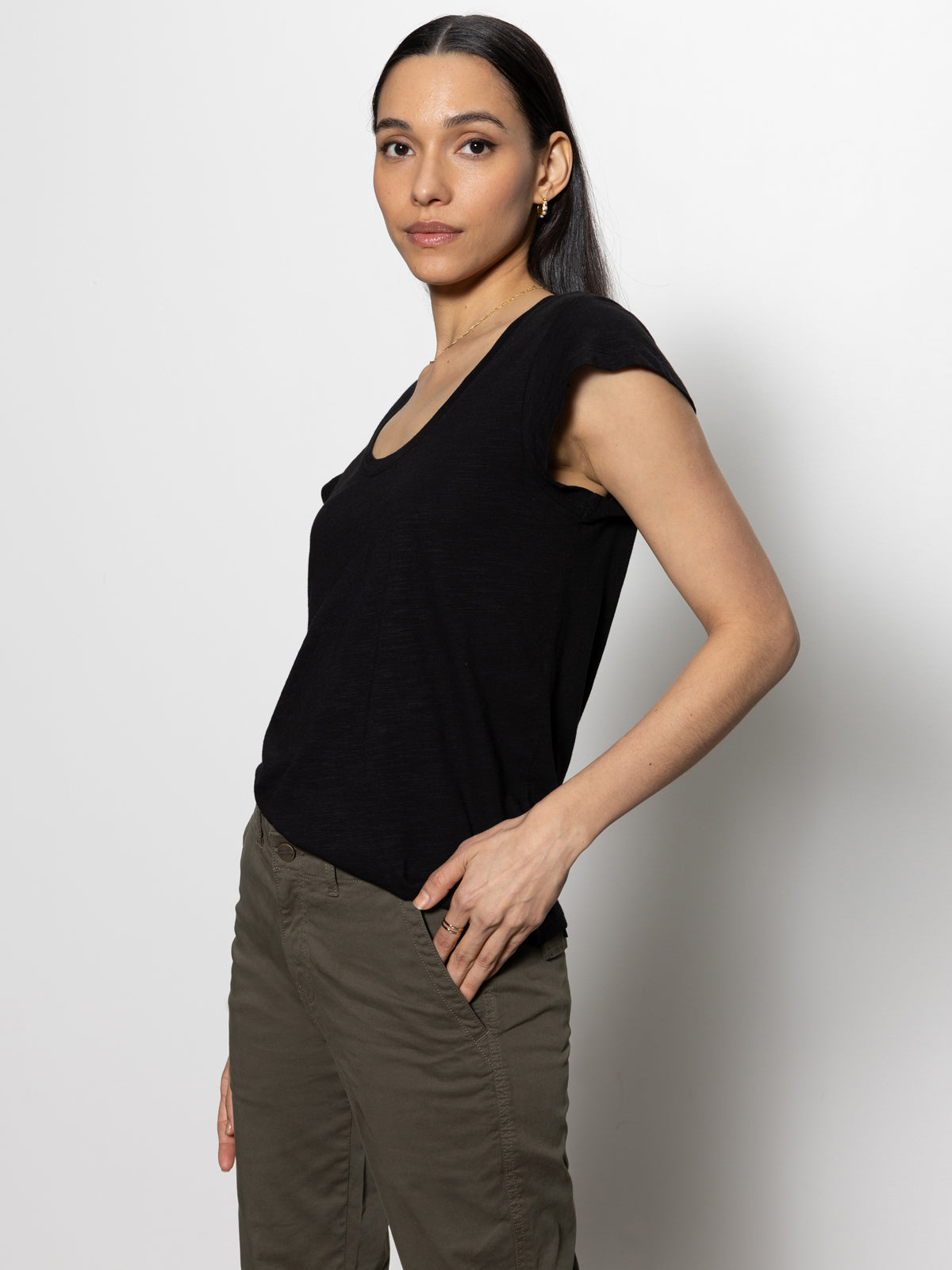 Against a plain light background, a dark-haired person poses slightly sideways with one hand on their hip. They look towards the camera while wearing Sanctuary Clothing's West Side Tee in black paired with olive green pants.