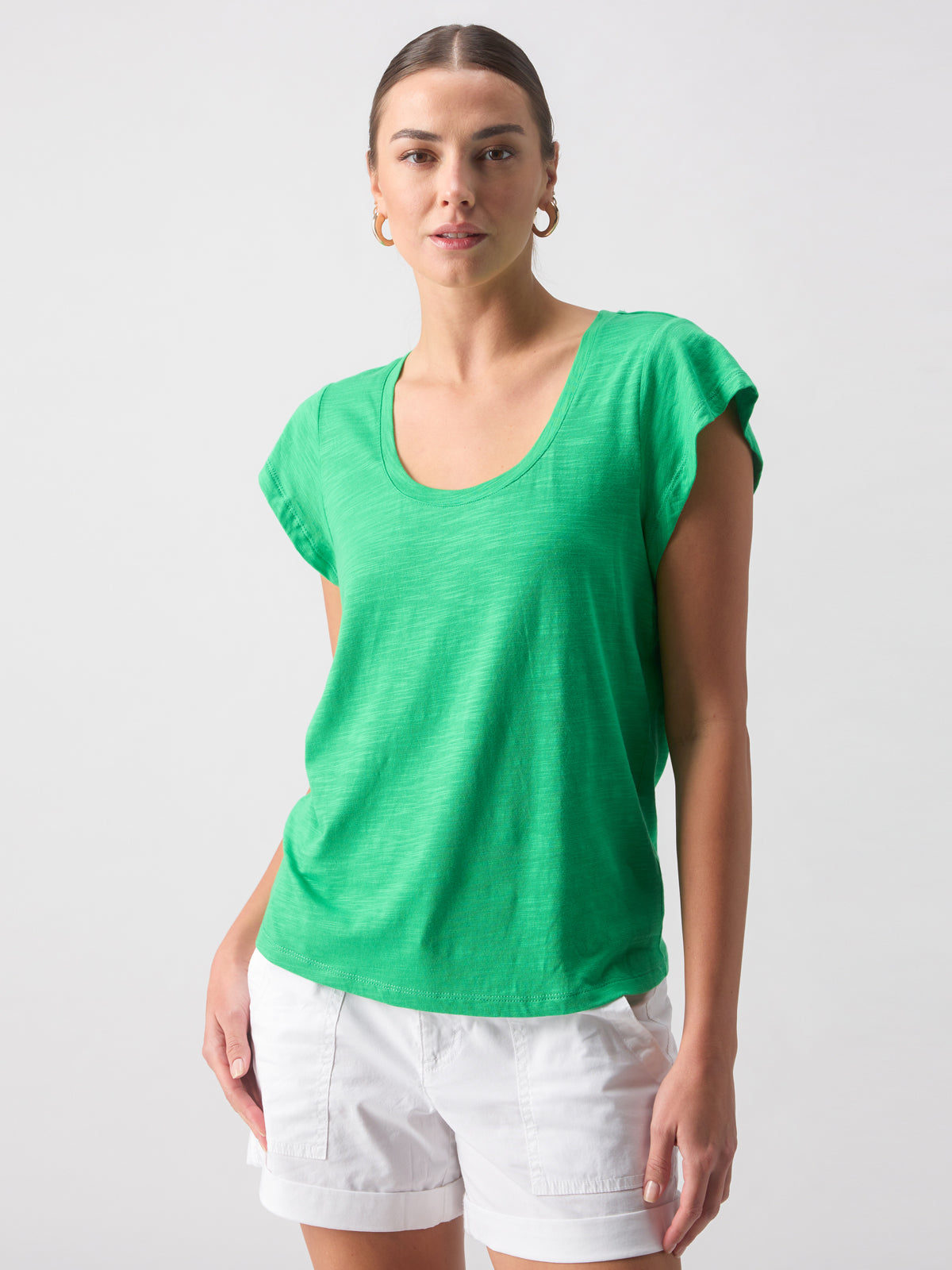 A person with light skin and brown hair pulled back is wearing the Sanctuary Clothing West Side Tee Green Goddess and white shorts. They are standing against a plain white background, looking directly at the camera with a slight smile. The individual is also wearing gold hoop earrings.