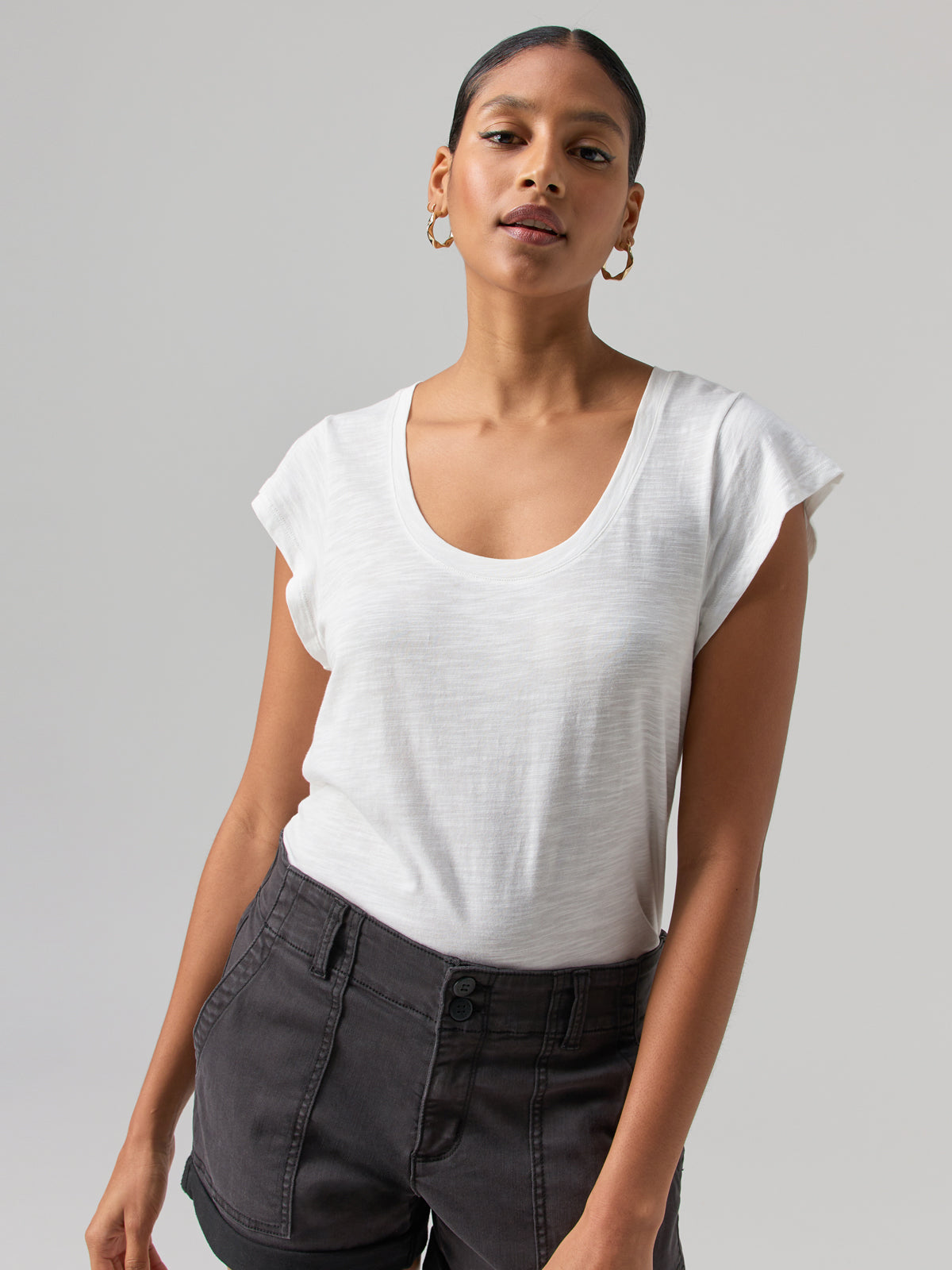 A person with short hair is wearing the West Side Tee in white from Sanctuary Clothing and dark gray high-waisted pants. They have gold hoop earrings and are standing against a plain background, looking directly at the camera with a slight smile.