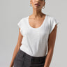 A person with short hair is wearing the West Side Tee in white from Sanctuary Clothing and dark gray high-waisted pants. They have gold hoop earrings and are standing against a plain background, looking directly at the camera with a slight smile.