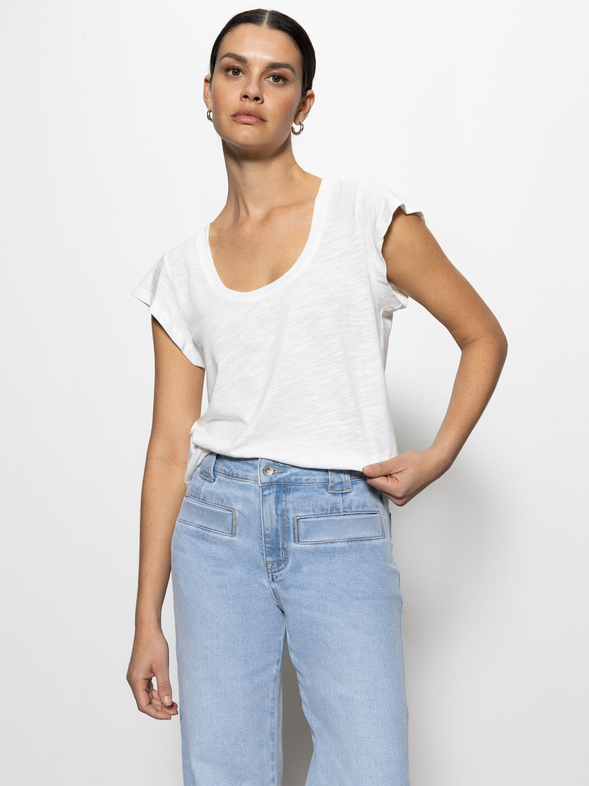 A person with dark hair is wearing a white scoop-neck T-shirt and light blue jeans. They have their hand on their hip and stand against a plain white background.