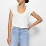 Wearing a Sanctuary Clothing 'West Side Tee' in white and light blue jeans, a person with dark hair tied back stands against a white background, with one hand on their hip.