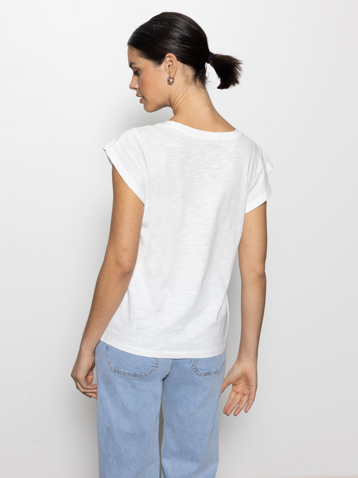 A woman with dark hair in a ponytail wears the Sanctuary Clothing west side tee in white and light blue jeans. Standing against a white background, she faces away from the camera, with one arm slightly bent.