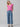 A person stands against a plain background, wearing a Sanctuary Clothing West Side Tee in Wild Pink with blue wide-legged cargo jeans, their hands in their pockets. They have long, light hair and are wearing sandals. Their expression is neutral, and their head is slightly tilted to one side.