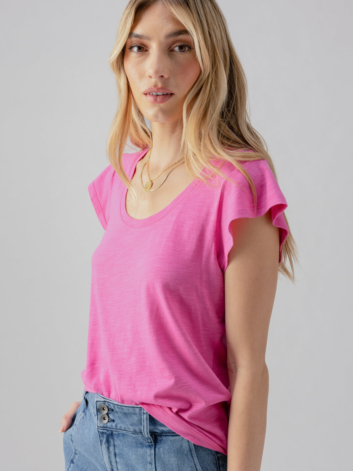 A person with long, blonde hair is wearing a Sanctuary Clothing West Side Tee in Wild Pink and light blue jeans. They are standing against a neutral background, with one hand placed in their pocket, looking towards the camera with a relaxed expression.