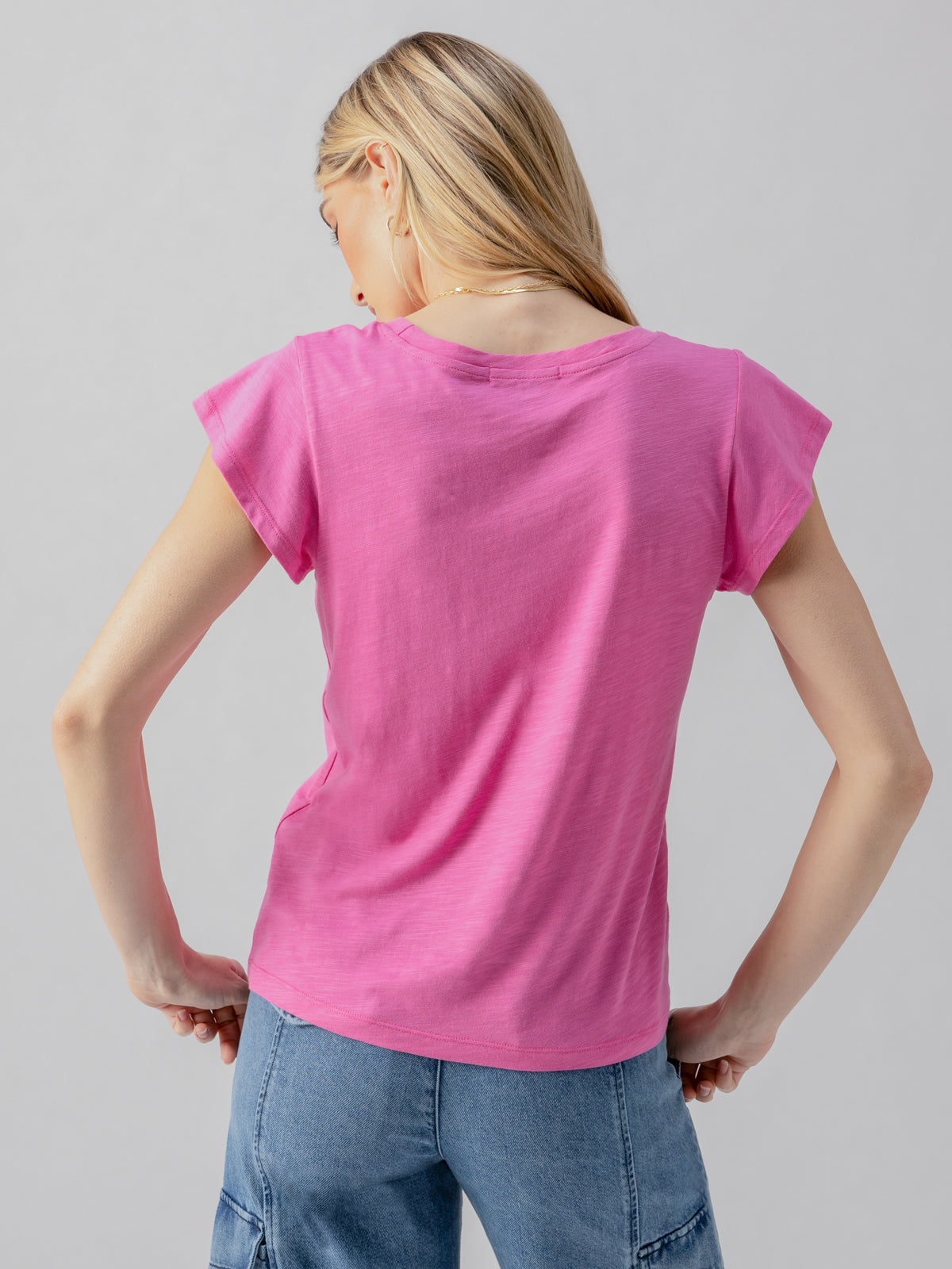 A person with long blonde hair is standing with their back to the camera, wearing a West Side Tee in Wild Pink by Sanctuary Clothing and light blue pants. The background is a plain, light grey color.