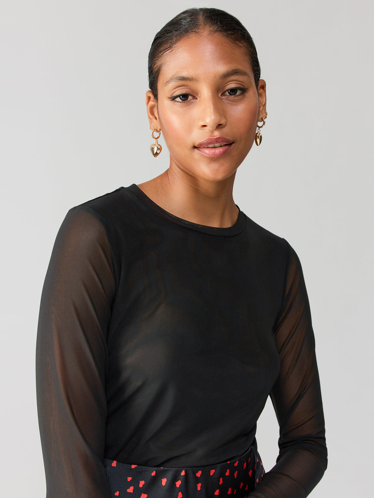 A woman with smooth, pulled-back hair looks at the camera. She is wearing the Main Squeeze Mesh Top Black by Sanctuary Clothing and gold hoop earrings. A subtle smile is on her face, and her skirt is patterned with small red shapes. The background is plain light gray.