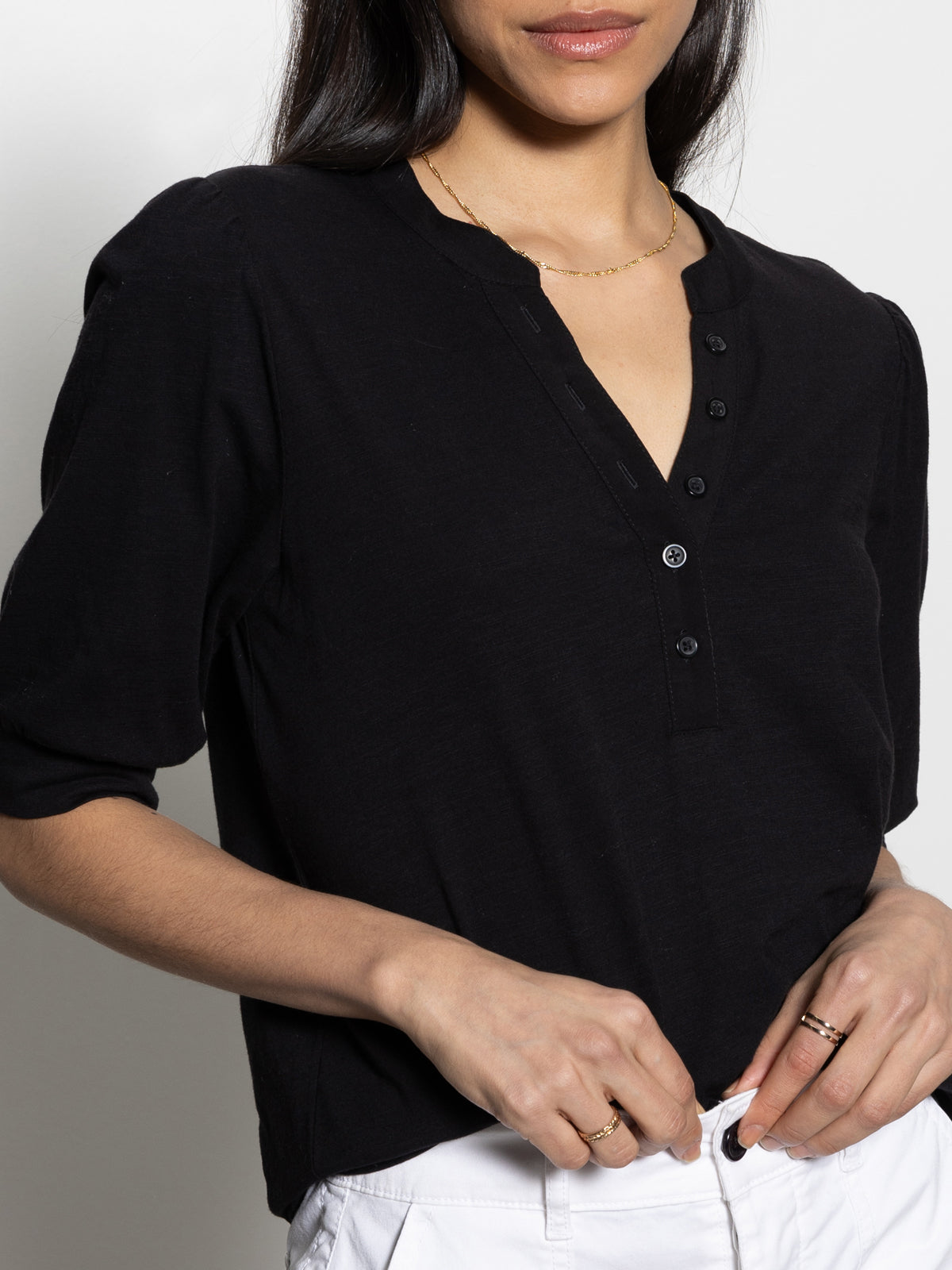 The image depicts a person wearing a Sanctuary Clothing mixed media tee in black, shown from shoulders to waist. With long dark hair, they touch their waistband while showcasing the textured fabric and short sleeves of the top alongside white pants.