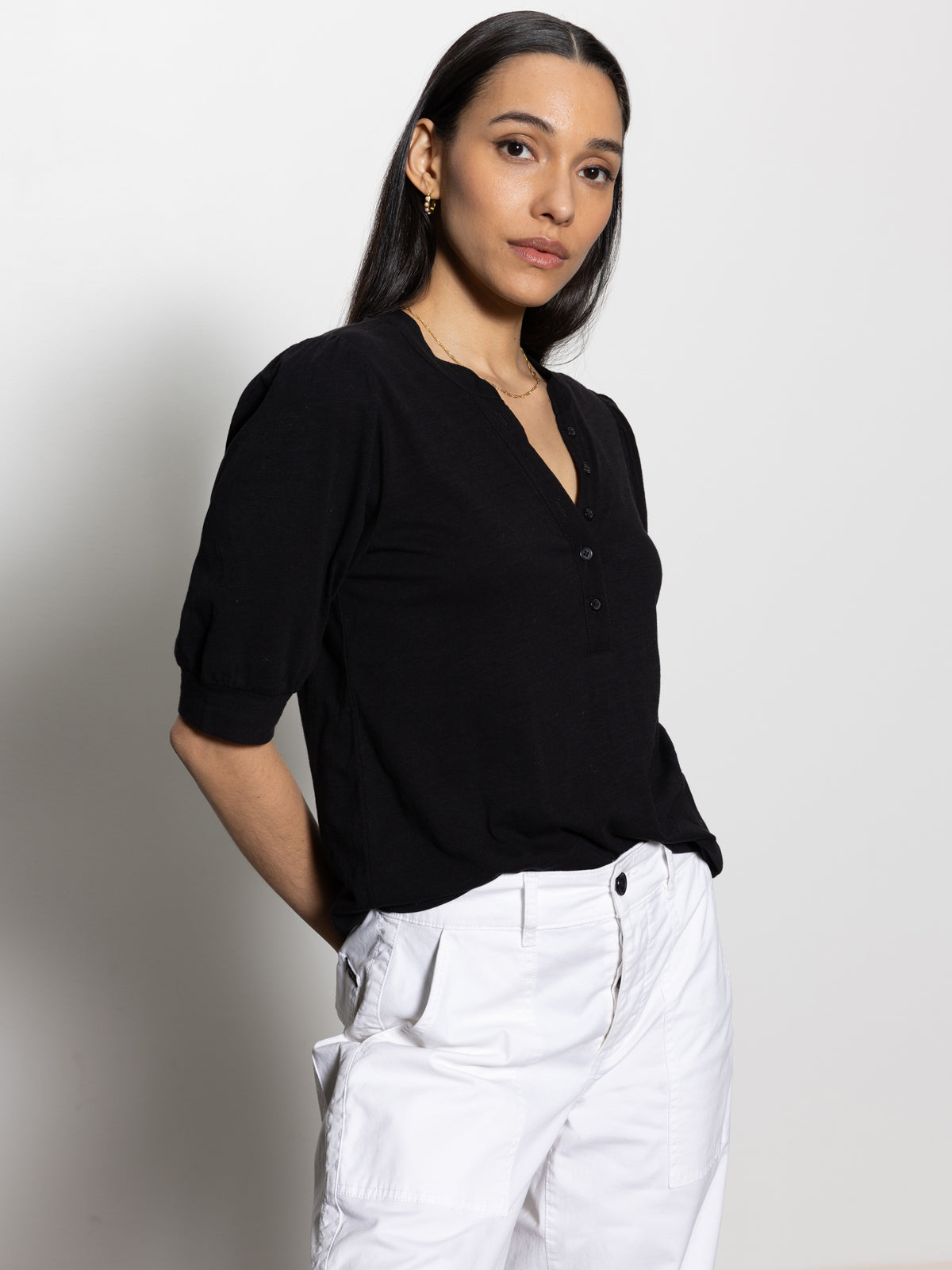 A person with long dark hair wears the Sanctuary Clothing mixed media tee in black paired with white pants. They're standing confidently against a plain light gray background, their hands resting loosely in their pockets.