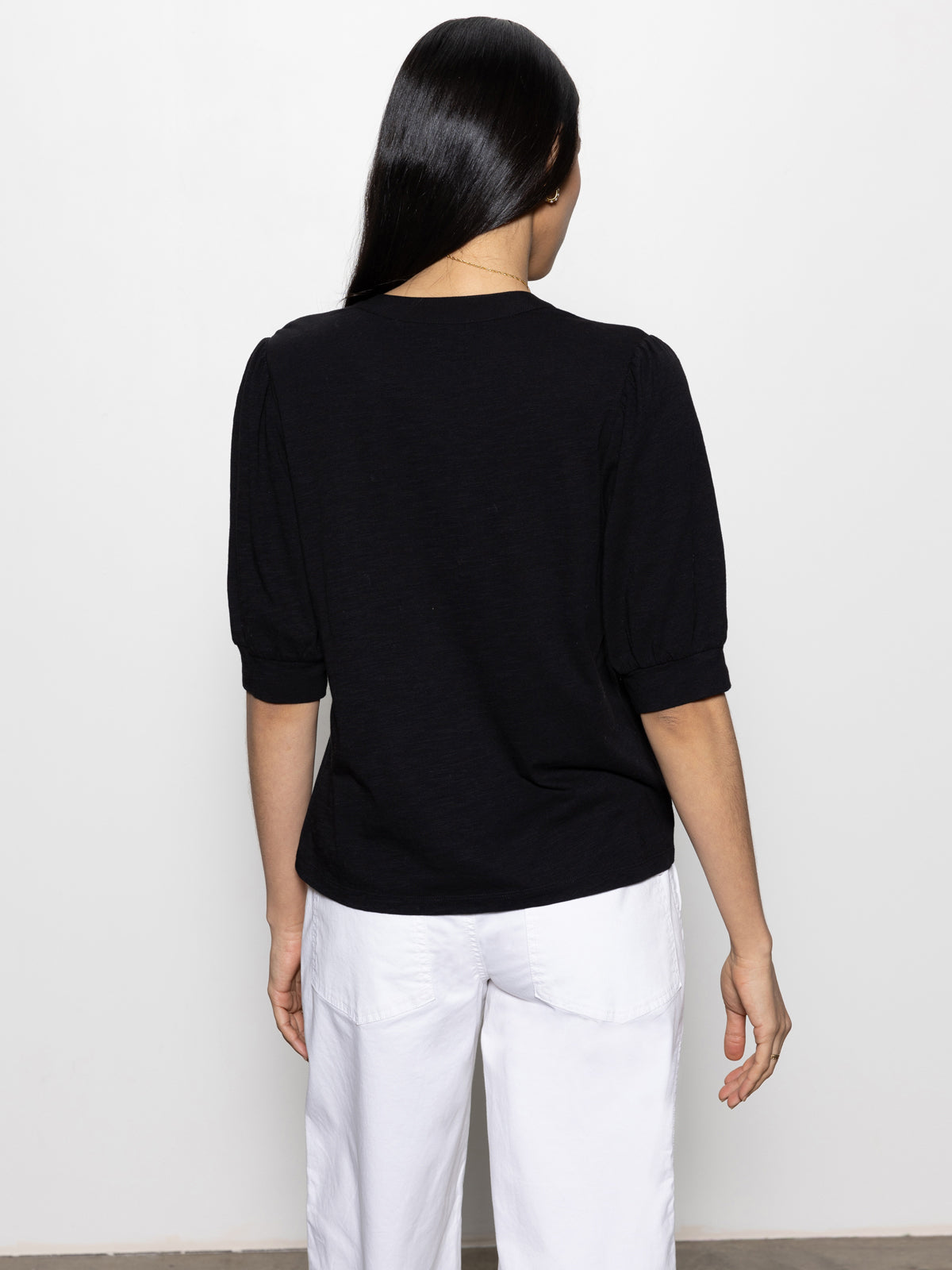 A woman with long black hair, standing with her back to the camera, is wearing a Sanctuary Clothing mixed media tee in black paired with white pants against a plain white background.