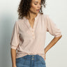 A woman with curly hair in a Sanctuary Clothing mixed media tee earth pink and blue jeans stands against a neutral background, one hand on her hip, gazing to the side.