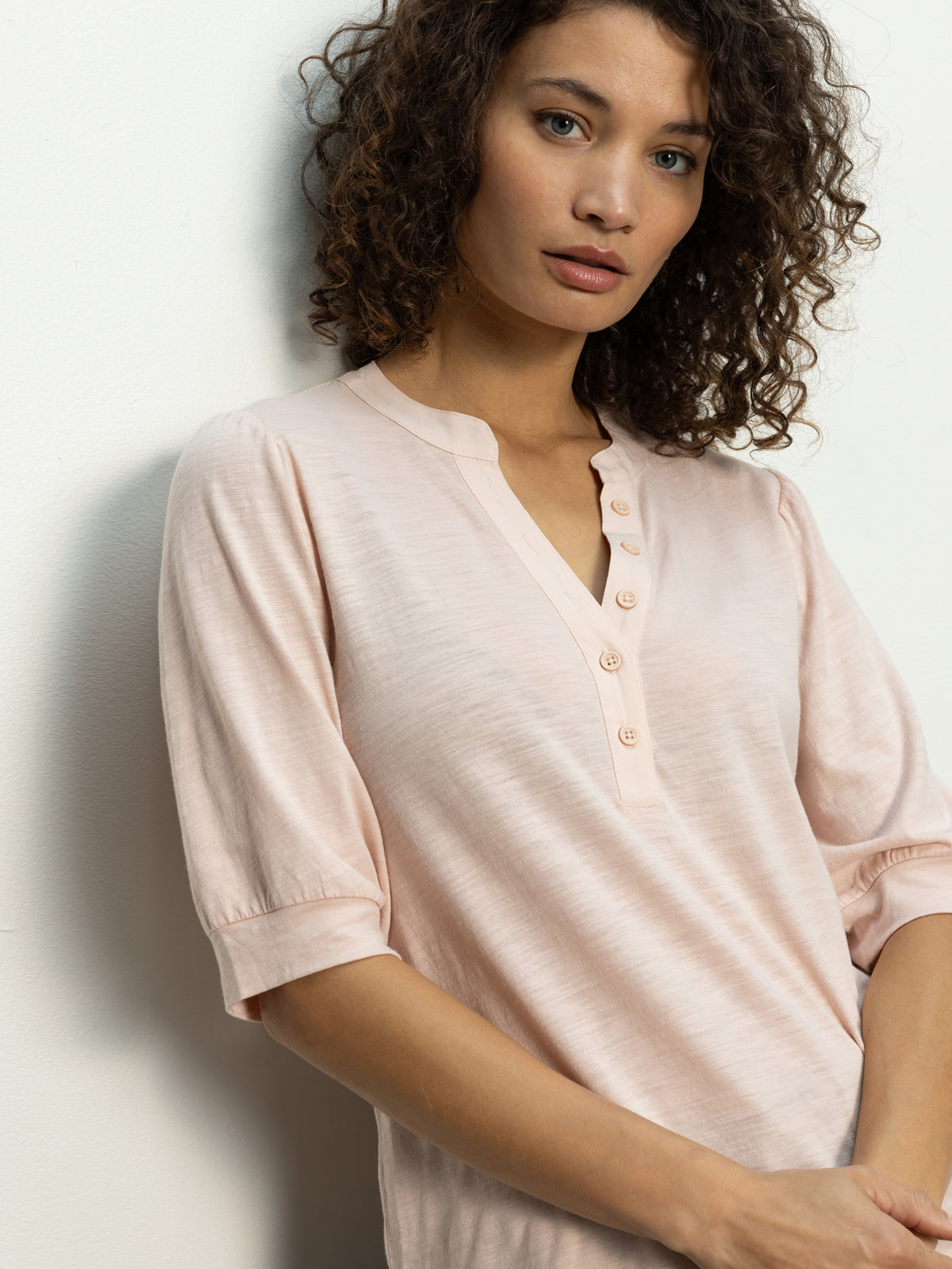 A person with curly hair leans against a white wall, wearing a Sanctuary Clothing mixed media tee in earth pink. They have a neutral expression and look towards the camera.