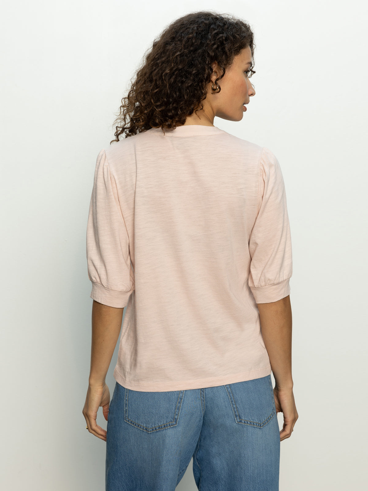 A person with curly hair stands with their back to the camera, wearing Sanctuary Clothing's mixed media tee in earth pink, featuring puffed three-quarter sleeves, and blue jeans. The background is a plain, light-colored wall.