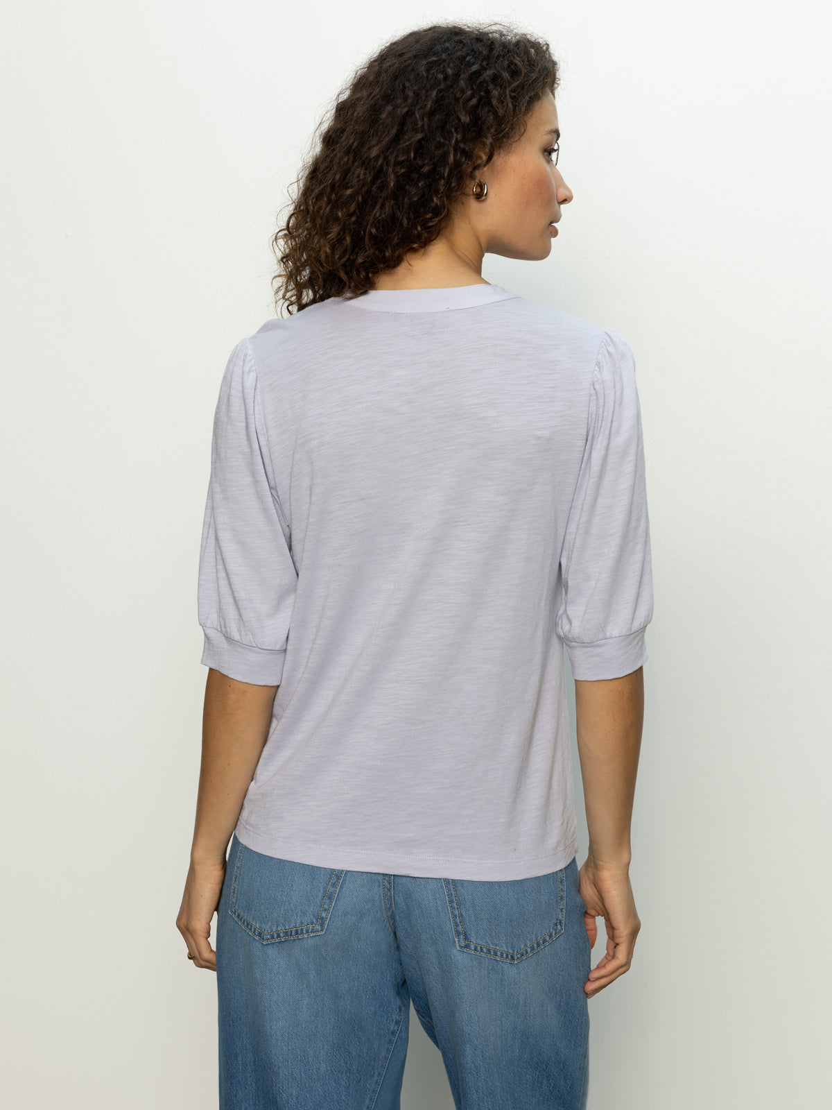 A person with curly hair stands with their back facing the camera, wearing a mixed media tee in lavender mist by Sanctuary Clothing and blue jeans, against a plain off-white background.