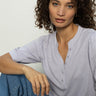 A person with curly hair sits casually, wearing a Sanctuary Clothing mixed media tee in lavender mist and blue jeans. They exude a relaxed, thoughtful expression while looking towards the camera against a plain background.