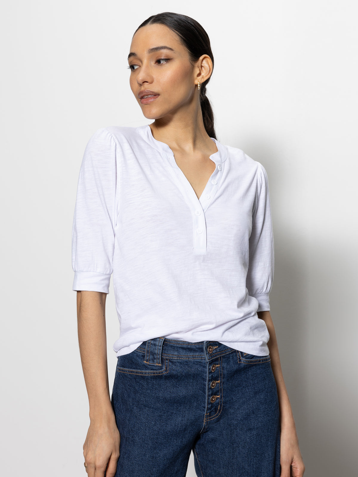 A person with dark hair tied back is wearing a mixed media tee white by Sanctuary Clothing and high-waisted blue jeans, standing against a plain white background, looking to the side with a calm expression.